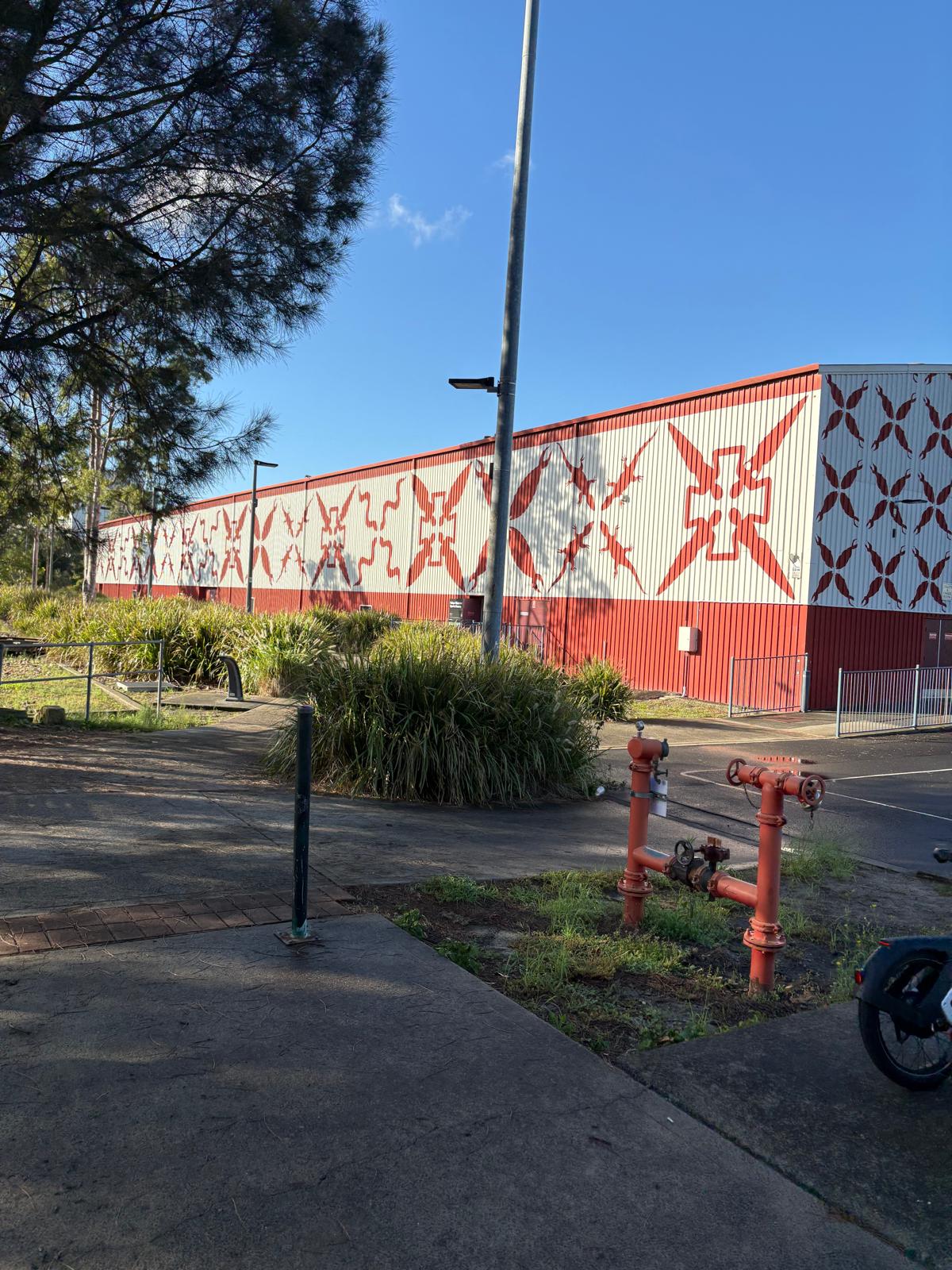 Industrial building with long white wall mural featuring red X-like shapes, red base, blue sky, and surrounding greenery and pipes nearby at ground level.