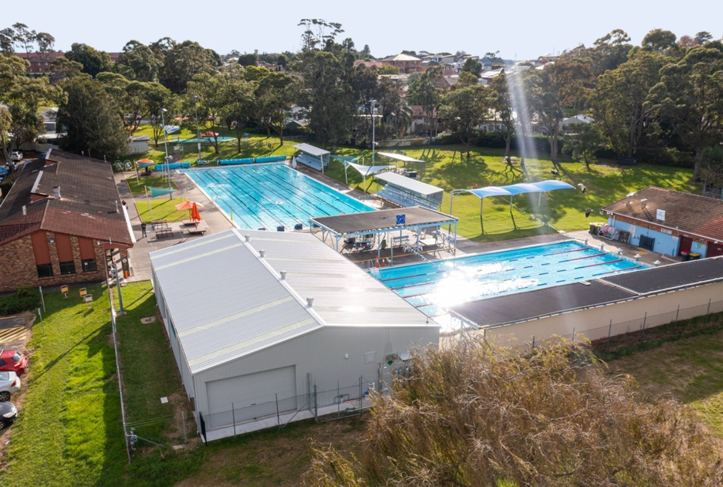 Aerial view of a large outdoor swimming complex with multiple blue pools, surrounding buildings, and trees, with a fountain shooting water into the air.